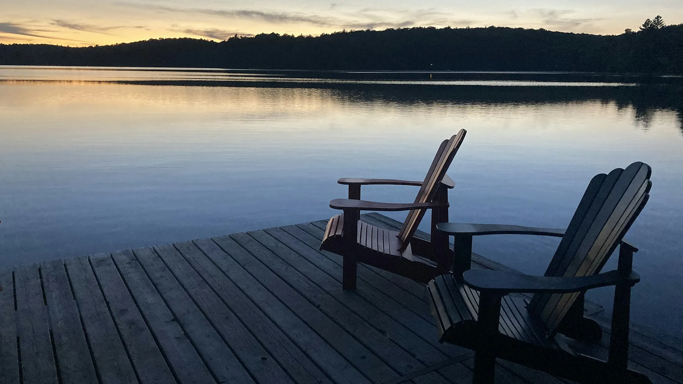 Two Muskoka chairs on a dock overlooking a calm lake at dusk